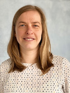 Brankatschk mit mittellangem blondem Haar, gepunkteter Bluse und hellem Hintergrund.